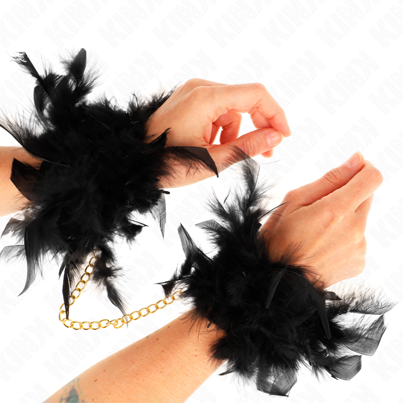 Detalle de las esposas de plumas negras Kink con cadena dorada, puestas en las muñecas.