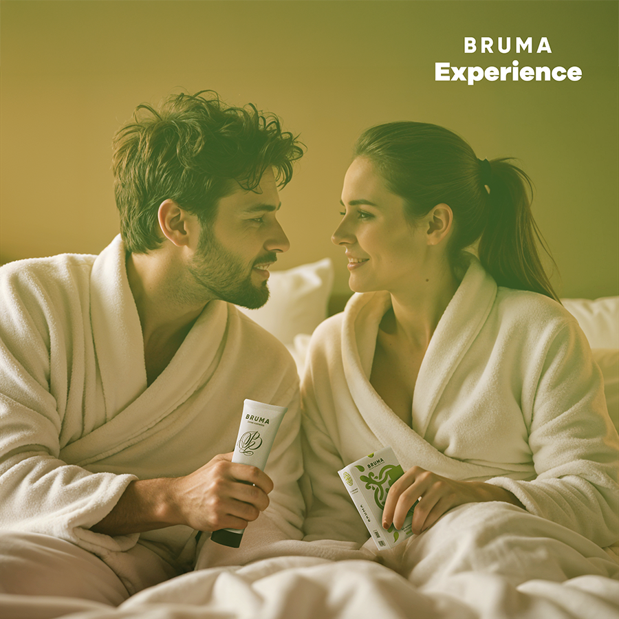 Pareja en albornoces blancos sosteniendo tubos de BRUMA Experience en la cama.